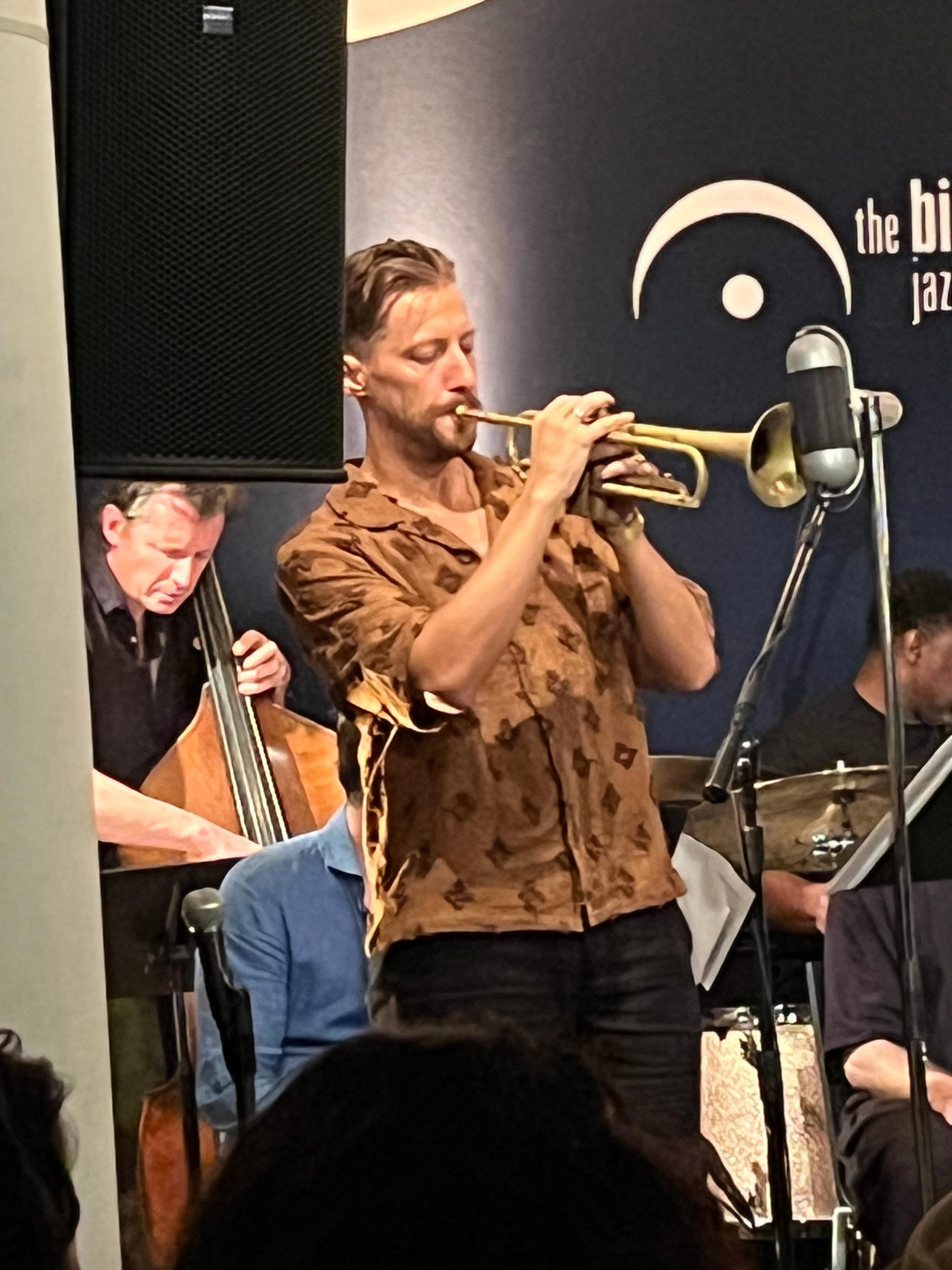 A man in an orange shirt plays the trumpet with a bassist and a drummer in the background