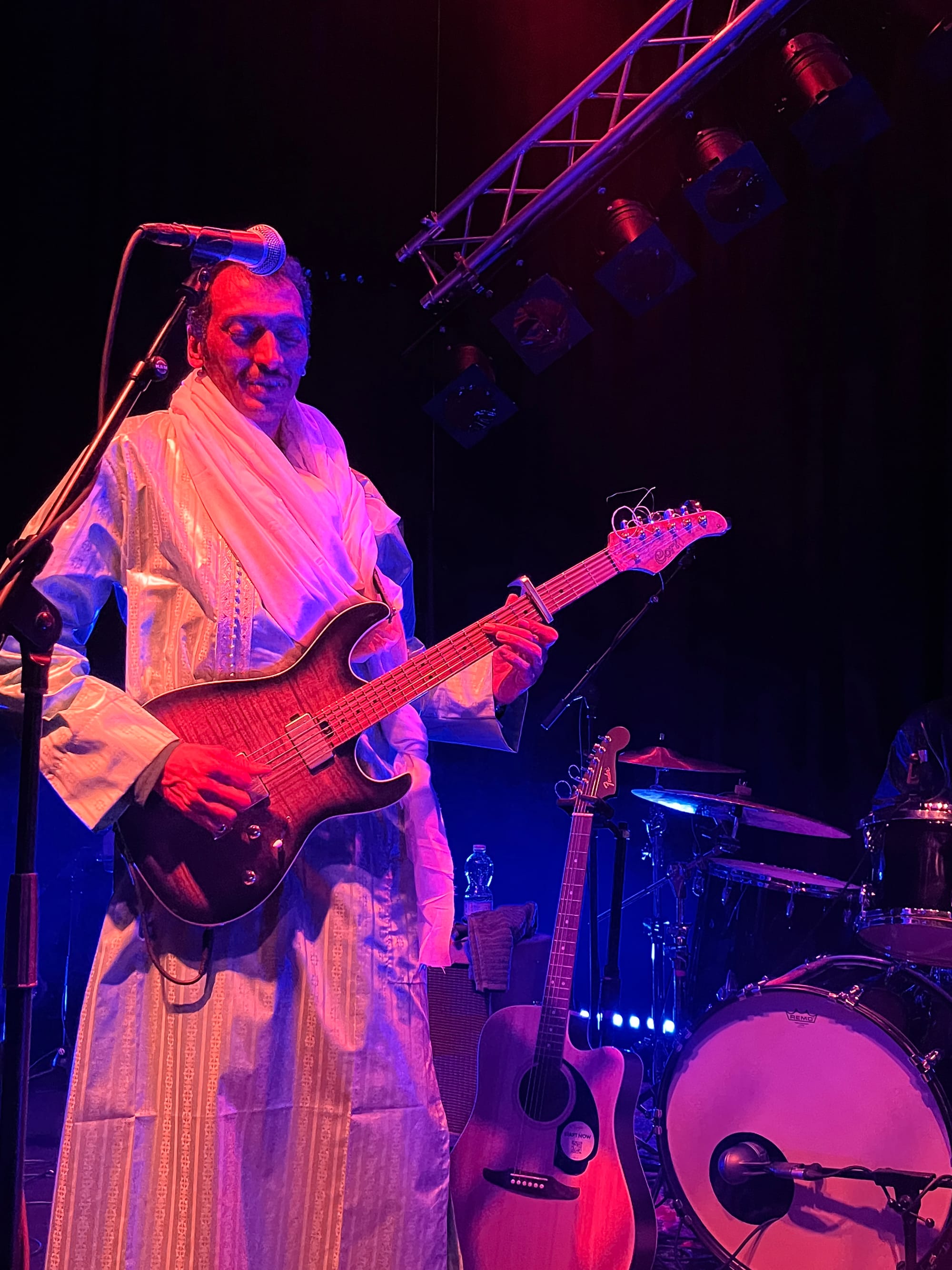 Bambino, in Tuareg dress, plays his electric guitar in the stage light at Sudhaus Basel, with his acoustic guitar behind him beside the drummer’s bass drum.